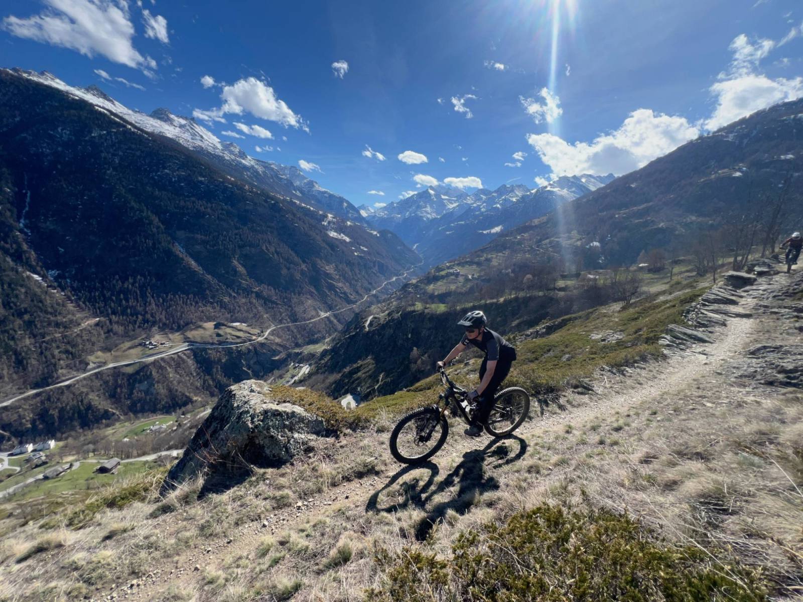 Mountain biker on a traversing trail above a Valais valley, snow-capped peaks behind