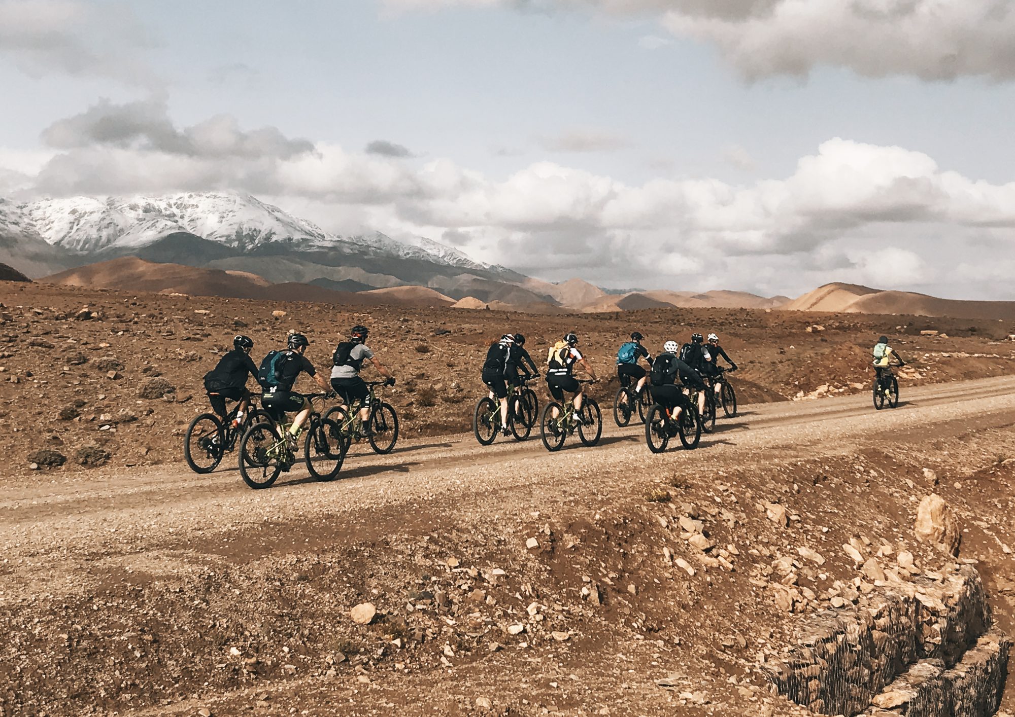 Eine Gruppe von Bikern mit MTB und E-Bike auf einer breiten Piste vor den verschneiten Gipfeln des Atlas, Reise Zagoride in Marokko mit Exoride